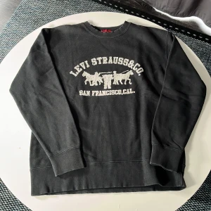 Svart sweatshirt från Levi's - Svart sweatshirt från Levi's med klassiskt tryck framtill i vitt med texten 'Levi Strauss & Co. San Francisco, Cal.' och ikoniska hästmotivet. Tröjan har rund hals, ribbade muddar och är tillverkad i mjuk bomull. Perfekt för en avslappnad och cool stil.
