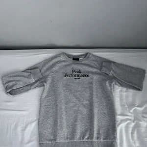 Grå sweatshirt från Peak Performance - Säljer en stilren grå sweatshirt från Peak Performance med svart logga tryckt på bröstet. Tröjan har rund halsringning, ribbade muddar och är tillverkad i mjuk bomullsmix. Perfekt för en avslappnad och sportig look.