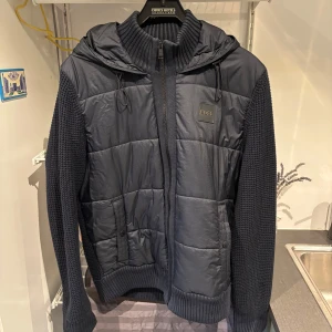 HUGO BOSS Cardigan Jacka | Mörkblå Strl M - Säljer en mörkblå cardigan från HUGO BOSS med stickade ärmar och quiltad front. Tröjan har huva med snörning, dragkedja och ribbade muddar. BOSS-logga på bröstet och praktiska fickor framtill. Den är använd men har inga skador överhuvudtaget och är i fortsatt mycket gott skick! 