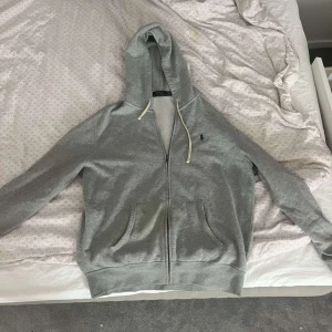 Grå hoodie från Polo Ralph Lauren - Snygg grå hoodie från Polo Ralph Lauren med dragkedja framtill, huva med vita snören och två fickor. Klassisk broderad logga på bröstet. Perfekt för chill dagar och enkel att matcha med jeans eller joggers.