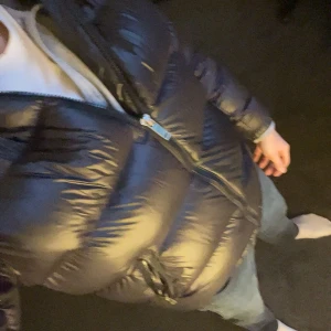 Svart pufferjacka med huva - Säljer en svart pufferjacka med glansig finish och huva. Jackan har dragkedja framtill och två fickor med dragkedja på sidorna. Den är vadderad och har en klassisk pufferlook som håller dig varm under kalla dagar.