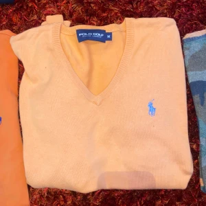 Beige v-ringad tröja Polo Golf - Snygg beige v-ringad tröja från Polo Golf Ralph Lauren med blå broderad logga på bröstet. Tröjan är i mjuk bomull och har ribbade muddar vid hals, ärmslut och nederkant. Perfekt för en stilren och klassisk look.