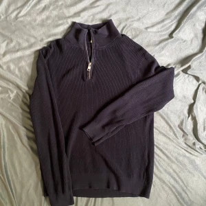 Svart stickad tröja med half zip - Mörk, stickad tröja i svart med hög krage och half zip-dragkedja framtill. Tröjan har lång ärm och ribbad struktur, perfekt för lager-på-lager under kyliga dagar.