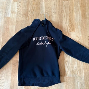 Mörkblå Burberry hoodie med tryck - Snygg mörkblå hoodie från Burberry med vit text 'Burberry London, England' på bröstet. Klassisk huva med snörning och långa ärmar. Perfekt för dig som gillar streetstyle och vill ha något trendigt och bekvämt.