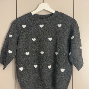 Grå stickad tröja med hjärtan Vero Moda - Supermysig grå stickad tröja från Vero Moda med vita hjärtan över hela. Tröjan har rund halsringning och lite lösare passform, perfekt för kyliga dagar. Snygg att styla med jeans eller kjol för en söt look.