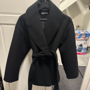 Svart kappa med bälte från Gina Tricot - Snygg svart kappa från Gina Tricot i storlek XS. Modellen har bred krage och knyts med ett bälte i midjan för en clean och stilren look. Perfekt för dig som gillar klassiska vibes och vill ha en jacka som funkar till många outfits.