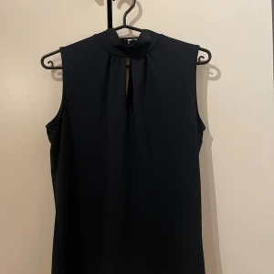 Svart ärmlös blus från Stockh LM - Snygg svart blus från Stockh LM i storlek XS. Modellen är ärmlös med hög hals och en liten öppning framtill som ger en cool detalj. Perfekt att styla med jeans eller kjol för en trendig look.