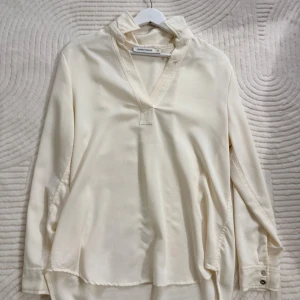 Krämvit henley från Carin Wester - Stilren krämvit henley-skjorta från Carin Wester med v-ringning och klassisk krage. Skjortan har långa ärmar med knappar vid manschetten och är tillverkad i ett mjukt, slätt material som ger en clean look.