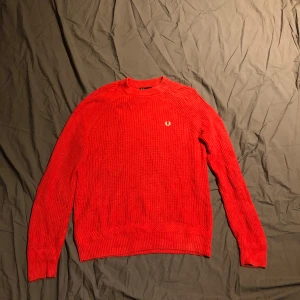 Röd stickad tröja Fred Perry M - Snygg röd stickad tröja från Fred Perry i storlek M. Tröjan har ribbad struktur, rund hals och det klassiska Fred Perry-logot på bröstet i vitt. Perfekt för dig som gillar stilrena och sportiga plagg med lite edge.