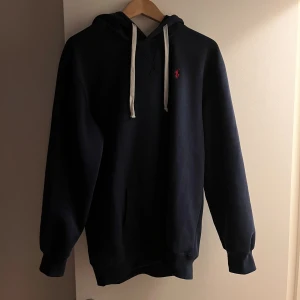 Mörkblå hoodie från Polo Ralph Lauren - Snygg mörkblå hoodie från Polo Ralph Lauren med vit snörning och klassisk röd logga på bröstet. Tröjan har huva, känguruficka och ribbade muddar. Perfekt för en chill och stilren look. Stl L