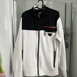 Svartvit vindjacka från Prada - Snygg svartvit vindjacka från Prada med dragkedjor och röda detaljer. Jackan har hög krage, lång ärm och flera fickor med zip. Klassisk logga på bröstet och sportig look som sticker ut.