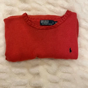 Röd stickad tröja från Polo Ralph Lauren - Snygg röd stickad tröja från Polo Ralph Lauren med klassisk rund hals och svart broderad logga på bröstet. Tröjan har långa ärmar och ribbade muddar vid hals, ärmslut och nederkant. Perfekt för dig som gillar stilrena och tidlösa plagg särskilt mysig inför vintern.  Pris kan diskuteras
