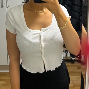 Vit croppad ribbad topp med knappar - Säljer en vit croppad topp med korta ärmar och ribbad struktur. Toppen har en snygg v-ringning och knappar framtill samt vågig kant nedtill. Perfekt för dig som gillar stilrena och trendiga plagg.
