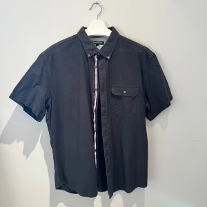 Svart kortärmad skjorta Banana Republic - Svart oxfordskjorta från Banana Republic i storlek XL. Skjortan har korta ärmar, klassisk krage med knappar och en bröstficka med lock. Materialet är mjukt bomullstyg och passformen är normal. Perfekt för dig som gillar stilrena och enkla plagg. Hör av er vid frågor