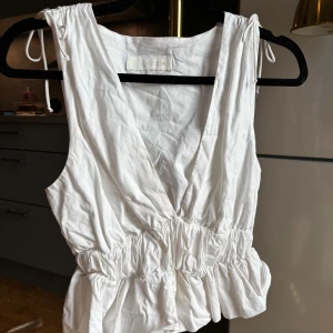 Vit omlottopp från Zara - Säljer en vit omlottopp från Zara med djup v-ringning och knytband på axlarna. Toppen har resår i midjan som ger en snygg volangkant nedtill. Perfekt för dig som gillar en luftig och trendig look.