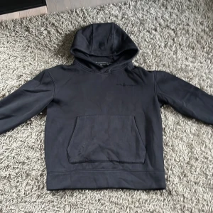 Svart hoodie från Sail Racing - Svart hoodie från Sail Racing med gummi logga på bröstet och emblem på huvan. Tröjan har en stor känguruficka framtill, mjuk fleece på insidan och långa ärmar. Perfekt för dig som gillar en stilren och sportig look.