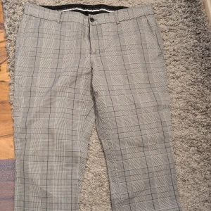 Grå rutiga kostymbyxor från Bläck - Snygga grå kostymbyxor från Bläck i klassiskt rutig mönster. Byxorna är tillverkade i 100% bomull och har raka ben, bälteshällor och fickor både fram och bak. Perfekta för en stilren look.