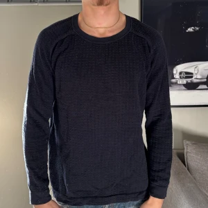 Sweatshirt - Säljer en mörkblå långärmad tröja från Filippa K med rund hals och ett snyggt strukturerat rutmönster i tyget. Tröjan har en normal passform och är tillverkad i mjuk bomull, perfekt för dig som gillar stilrena och bekväma plagg.