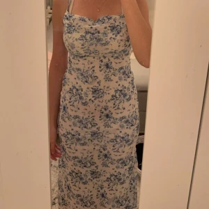 Blommig långklänning från SHEIN - Superfin långklänning från SHEIN med blått blommönster på vit botten. Klänningen har smala axelband, snörning i ryggen och en figurnära passform. Perfekt för sommarens alla tillfällen! Materialet är lätt och känns svalt mot huden.