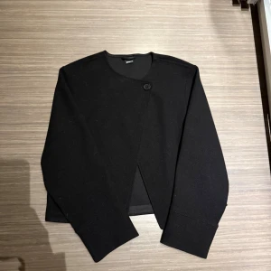 Svart cardigan jacka från Gina Tricot - Svart cardigan jacka från Gina Tricot i stilren design med rund hals och en knapp framtill. Jackan har lång ärm och är tillverkad i mjukt material som ger en clean och enkel look. Perfekt för dig som gillar minimalistisk stil.