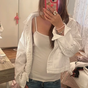 Vit jeansjacka med broderade detaljer - Säljer en vit jeansjacka från Dilvin Jeans med snygga broderade hålmönster på ärmar och framstycke. Jackan har klassisk krage, knappar framtill och två fickor. Perfekt för dig som vill ha en unik och fräsch look.