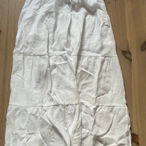 Vit långkjol i bomull - Luftig och flowy vit långkjol i bomull med flera volanger och lätt struktur i tyget. Perfekt för varma dagar och har en avslappnad vibe. Kjolen har en enkel design utan mönster och är lätt att matcha med olika toppar. Den har tyvärr en fläck på sig