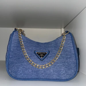 Blå axelväska i denim  - Snygg blå axelväska  i denim-look med guldfärgad kedja och klassisk trekantig logga framtill. Väskan har en mjuk, rundad form och detaljer i blått läder. Perfekt för dig som vill ha en trendig och lyxig accessoar.