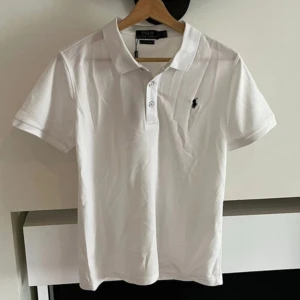 Vit Polo Ralph Lauren t-shirt slim fit - Vit t-shirt från Polo Ralph Lauren i slim fit-modell med klassisk krage och två knappar framtill. Ikoniska broderade loggan i svart på bröstet. Tillverkad i mjuk bomull som känns skön mot huden. Perfekt för dig som gillar stilren och enkel design.