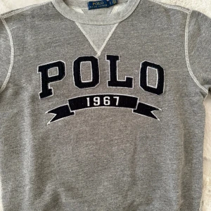 Grå sweatshirt från Polo Ralph Lauren - Snygg grå sweatshirt från Polo Ralph Lauren med stor svart och vit POLO 1967-logga framtill. Klassisk rund halsringning och mjukt material som är perfekt för chill dagar. Stilren och enkel att matcha med jeans eller joggers.