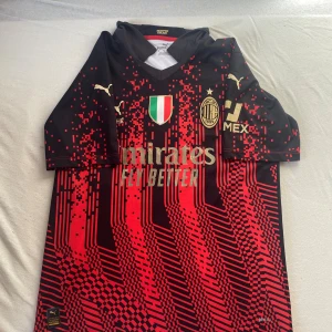 AC Milan Bennacer #4 Puma fotbollströja - AC Milan matchtröja med Bennacer och nummer 4 på ryggen. Svart och röd med digitalt mönster, klubbmärke och sponsortryck på bröstet. Tillverkad i lätt polyester med Drycell-teknologi för bästa komfort på planen. Klassisk V-ringning och korta ärmar.