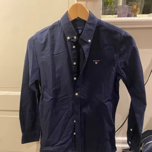 Marinblå skjorta från GANT - Snygg marinblå skjorta från GANT med klassisk button-down krage och vita knappar. Skjortan har lång ärm och broderad logga på bröstet. Tillverkad i mjuk bomull som känns skön mot huden. Perfekt för dig som gillar stilrena och tidlösa plagg.