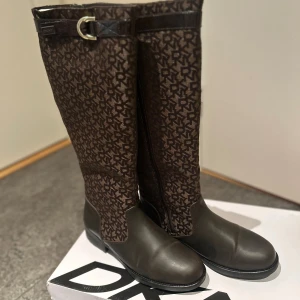 Bruna DKNY stövlar med monogram - Snygga höga stövlar från DKNY i mörkbrunt med all-over DKNY monogram. Stövlarna har rund tå, platt sula och dekorativt spänne upptill. Materialet är en mix av skinn och textil, vilket ger en lyxig känsla. Perfekt för dig som gillar statement pieces och designer.