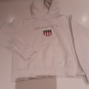 Vit hoodie från GANT med logga - Vit hoodie från GANT med broderad logga och sköld på bröstet. Klassisk känguruficka framtill, huva och långa ärmar. Perfekt för dig som gillar stilren och sportig look.