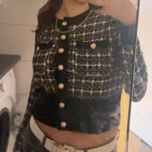 Rutig svartvit kofta med guldknappar - Snygg croppad kofta i svart och vitt rutigt mönster med dekorativa guldknappar framtill och på fickorna. Koftan har långa ärmar och en klassisk, elegant look som passar perfekt till jeans eller kjol.