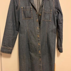 Lång jeansklänning från Lindex - Säljer en lång jeansklänning från Lindex i klassisk blå denim. Klänningen har långa ärmar, två bröstfickor med knappar och knäppning framtill. Perfekt för dig som gillar en avslappnad men snygg look.