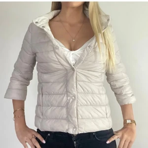 Beige dunjacka med knappar - Snygg beige dunjacka med quiltad design och tryckknappar framtill. Jackan har en lätt, puffig känsla och en feminin passform. Perfekt för dig som vill ha en stilren och enkel look under kyligare dagar.