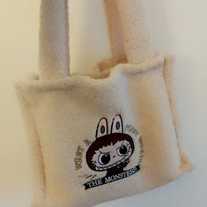 Beige fluffig axelväska med brodyr - Supermjuk och fluffig beige axelväska i teddy-material med breda handtag. På framsidan finns en broderad söt monsterfigur med texten 'THE MONSTERS'. Perfekt för dig som gillar gulliga och unika accessoarer.