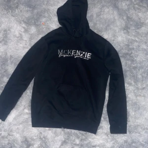 Svart hoodie från McKenzie - Svart hoodie från McKenzie med vit logotyp på bröstet. Tröjan har huva med snörning och en stor magficka framtill. Perfekt för chill dagar och enkel att matcha med jeans eller joggers. Mjuk och skön känsla tack vare bomullsmaterialet.