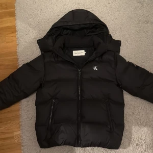 Svart pufferjacka från Calvin Klein Jeans - Hej! Jag säljer denna svarta pufferjacka från Calvin Klein Jeans med huva och dragkedja. Jackan har två sidofickor med dragkedja och tydliga CK-loggor på bröstet och ärmen. Vid armbågen så har det kommit ett litet hål, som går att sy väldigt enkelt och som inte syns så mycket vid användning. Jackan är väldigt varm och skön att ha på sig, den är väldigt skön och varm och fungerar perfekt till vintern. Jackan köptes för 2699, och säljs för endast 1799, priset kan kanske 💨lite vid snabbaffär endast