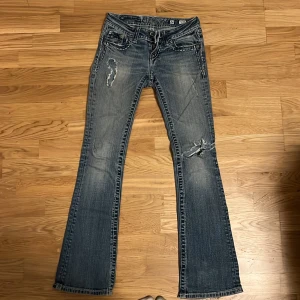 Miss Me bootcut jeans med broderi - Snygga ljusblå bootcut jeans från Miss Me med slitningar och broderade bakfickor dekorerade med pärlor och korsmönster. Jeansen har låg midja, klassisk femficksdesign och coola detaljer längs sömmarna. Köptes för 1200kr