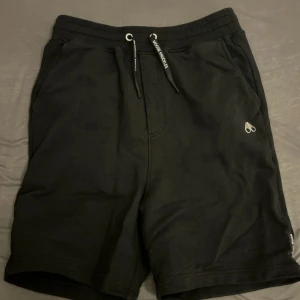 Svarta shorts från Moose Knuckles - Svarta shorts från Moose Knuckles med snygg logga på benet och snörning i midjan. Mjukt material och avslappnad passform, perfekta för chill dagar. Klassisk look med två sidofickor och elastisk midja.