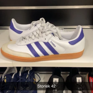 Adidas Samba vit/lila strl 42 - Säljer ett par klassiska Adidas Samba sneakers i vitt med lila detaljer och gummisula. Skorna har de ikoniska tre ränderna på sidan, snörning och låg profil. Perfekta för dig som gillar retrostil och sportig vibe.