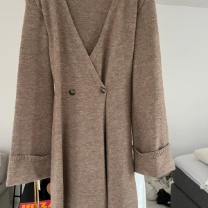 Adoore, beige stickad omlottklänning - Aldrig använd! beige stickad klänning med omlott-design och v-ringning. Klänningen har långa ärmar med uppvikta muddar och två knappar framtill. Perfekt för dig som gillar en avslappnad men ändå stilren look.