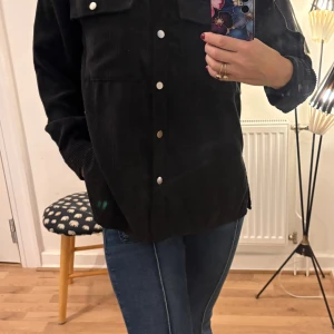 Svart manchester overshirt Gina Tricot - Svart overshirt i manchester från Gina Tricot med silvriga knappar framtill och på bröstfickorna. Skjortan har klassisk krage, långa ärmar och en loose passform. Perfekt att slänga över en t-shirt för en chill look.