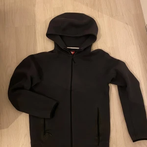 Äkta Svart hoodie från Nike med dragkedja - Priset är inte hugget i sten .Svart hoodie från Nike med hel dragkedja framtill och två fickor med dragkedja. Tröjan har huva och långa ärmar, perfekt för chill eller träning. Materialet är mjukt och känns som polyester. Enkel och stilren design utan synliga mönster.