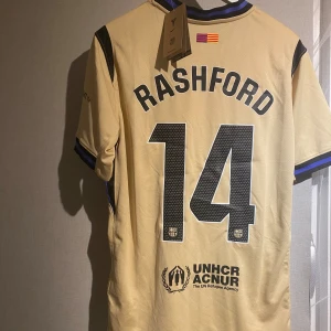 Barcelona Rashford #14 Nike fotbollströja - Exklusiv Barcelona fotbollströja med Rashford och nummer 14 på ryggen. Beige med svarta detaljer, FCB-emblem, Spotify-logga och UNHCR-tryck. Kortärmad med svart och blå krage, tillverkad i lätt polyester som andas. Perfekt för dig som vill sticka ut på planen.