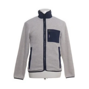 Grå fleecejacka Patagonia  XS - Grå fleecejacka från Patagonia i storlek XS. Jackan har mörkblå detaljer vid krage, fickor och bröstficka med dragkedja. Tillverkad av 100% återvunnen polyester och nylon. Perfekt för kyliga dagar med sin mjuka och sköna känsla.
