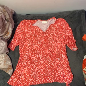 Röd blommig omlottklänning Holly & Whyte - Superfin röd kortklänning från Holly & Whyte med vitt blommönster och omlottmodell. Klänningen har korta ärmar och knytband i sidan för en snygg passform. Perfekt för sommarens alla tillfällen!