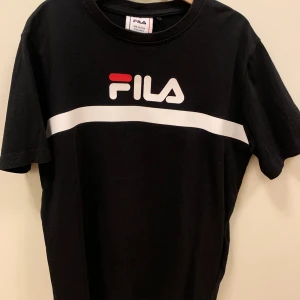 Svart FILA t-shirt med vit rand, stl 146/152 - Svart t-shirt från FILA med klassisk logga framtill och en vit horisontell rand över bröstet. T-shirten har rund halsringning och korta ärmar. Tillverkad i mjuk bomull för en skön känsla och avslappnad passform.