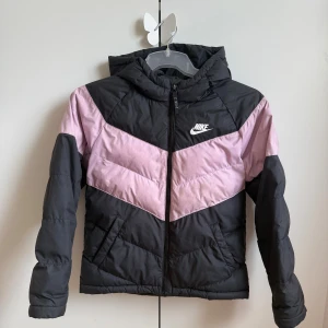 Svart och rosa pufferjacka från Nike - Nike pufferjacka i svart och ljusrosa med coolt V-mönster över bröst och rygg. Jackan har huva, dragkedja framtill och två sidofickor. Perfekt för kalla dagar och har en sportig vibe som passar streetstyle. Materialet är syntet och jackan är quiltad för extra värme ( jackan är barn M alltså typ 137-147 cm)